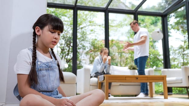 Stressed and unhappy young girl huddle in corner, cover her ears blocking sound of her parent arguing in background. Domestic violence at home and traumatic childhood develop to depression. Synchronos