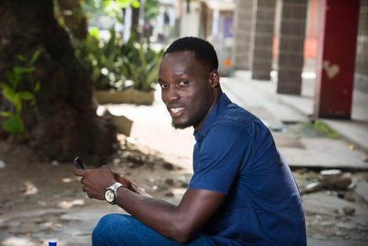 young man sitting outdoors in blue shirt looking at camera smiling with mobile phone.