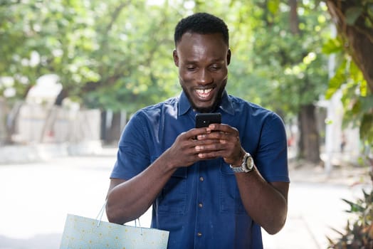 young man standing outdoors with shopping bag going to look at mobile phone while smiling.