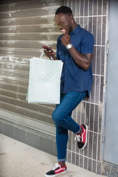 young man standing near a wall after shopping looking at mobile phone while smiling.
