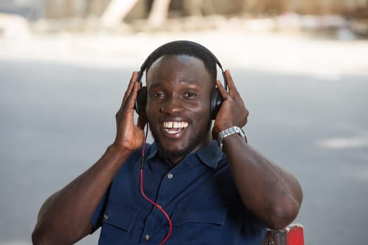 young man sitting outdoors listening to music using a headset and looking at camera smiling.