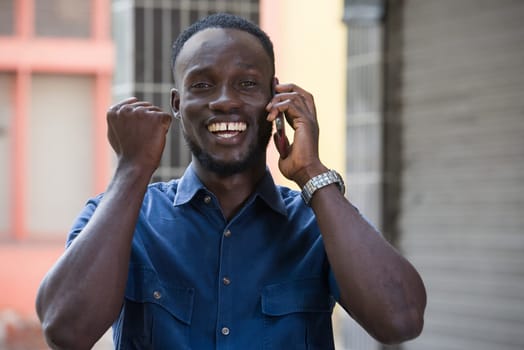 Photo of handsome excited man expressing surprise on face and gesturing while talking on the phone standing outside