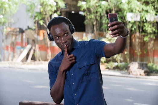 Man listens to music with headphones using a cell phone and makes photos with gesture of silence on the outside mouth.