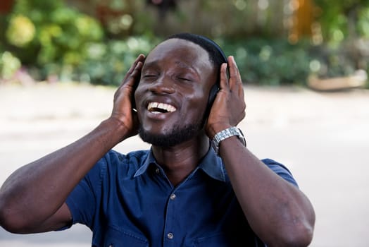 Young man standing outdoors listening to music using headphones while laughing.