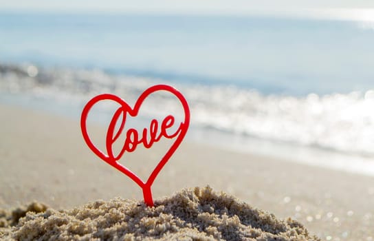 Plastic stick in shape of red heart and word Love in sand on beach seashore on sunny summer day close-up. Figures in shape of heart word Love on background sea waves. Love relationship romance concept