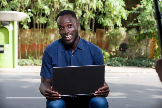 young man sitting outdoors with laptop watching camera smiling.