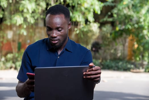 young man sitting outdoors with laptop watching mobile phone while smiling.