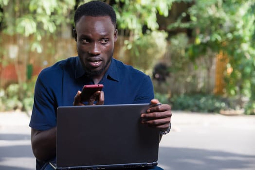 young man sitting outdoors with laptop computer communicating on cell phone while smiling.