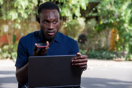 young man sitting outdoors with laptop computer communicating on cellphone.