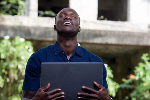 young man sitting outdoors smiling with his eyes closed with laptop.