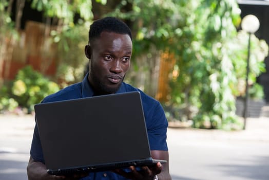 young man sitting outdoors thinking with laptop in hand while smiling.