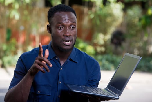 young man sitting outdoors talking with laptop in hand.