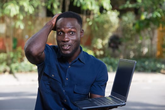 young man with laptop sitting outdoors looking at camera smiling.