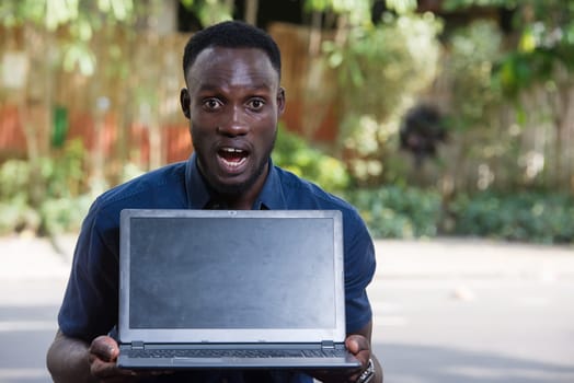 young man with laptop sitting outdoors looking at the camera looking surprised.