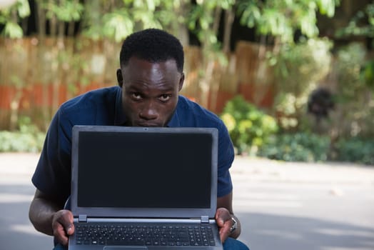 young man sitting outdoors holding a laptop watching the camera.