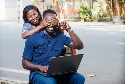 young man sitting outdoors working with laptop while his girlfriend covers his eyes with his hands smiling