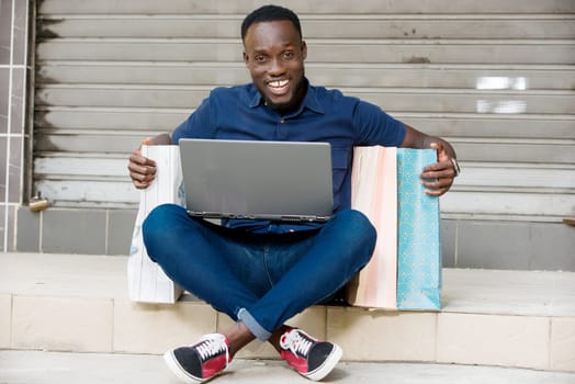 young man sitting on the floor with shopping bags and laptop looking at camera laughing.