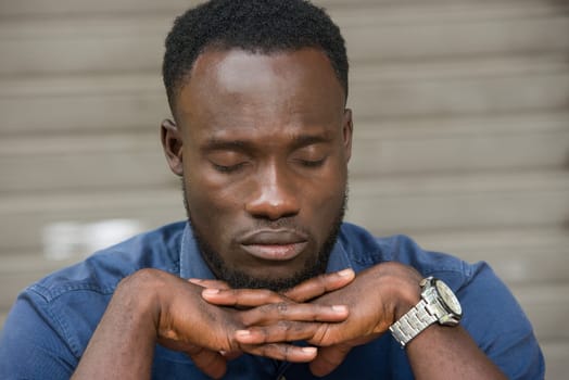young man in blue shirt sitting outdoors thinking of closed eyes.