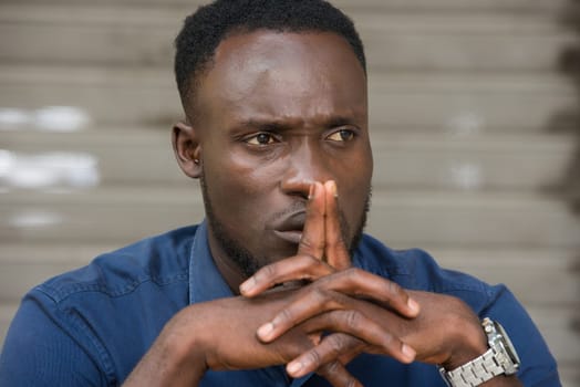 young man in blue shirt sitting outdoors thinking with fingers on his mouth.