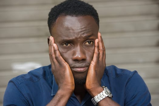 young man in blue shirt sitting outdoors thinking of hands on cheeks.