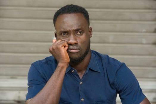 young man in blue shirt sitting outdoors thinking of fist under cheek.