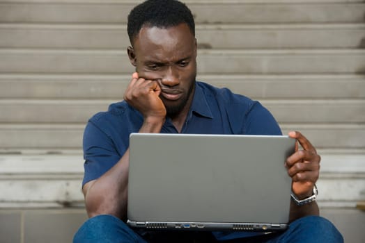 young man in blue shirt sitting outdoors looking at dissatisfied laptop.