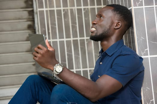 young man in blue shirt sitting near a wall thinking with laptop in hand.