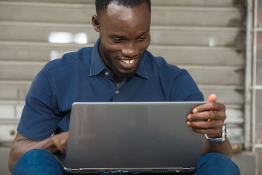 young man in blue shirt sitting outdoors looking at laptop smiling.