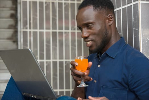 young man in blue shirt sitting near a wall looking at laptop smiling with a glass of juice in hand.