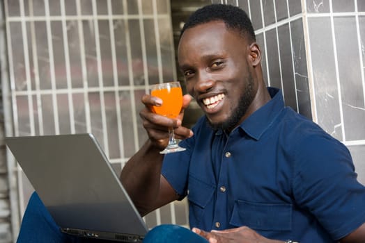 young man in blue shirt sitting near a wall looking at laptop smiling with a glass of juice in hand.