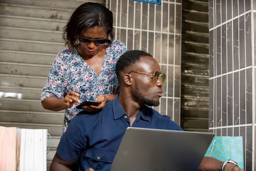 young couple sitting with shopping bags and looking at or using a laptop to make purchases online. woman standing behind her husband uses a mobile phone