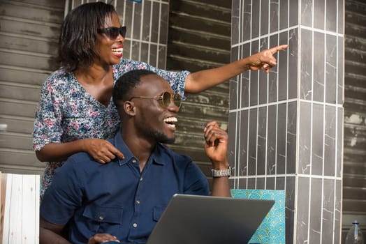 young couple sitting with shopping bags and using a laptop to make online purchases. couple laugh and look aside, woman pointing away