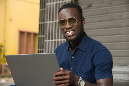 young man sitting outside with laptop watching the camera smiling.