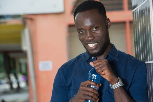 young man standing near a wall opening a bottle of water smiling.