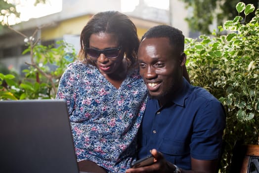 young couple sitting in a park looking at laptop smiling.