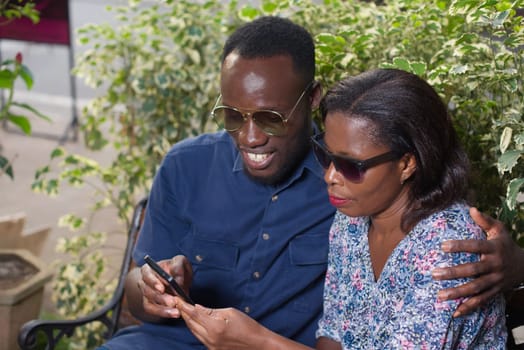 young couple sitting in a park looking at mobile phone while smiling.