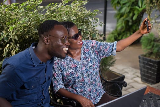 young couple sitting in a park in sunglasses photographing themselves with mobile phone while smiling.