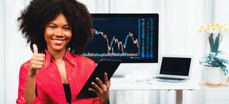 Analytical young African American businesswoman, a specialist in successful stock exchange trading, against dynamic data graph displaying marketing trend analysis on screen. Tastemaker.