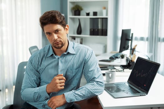 Profile of smart investor stock in trader looking at camera in dynamic exchange on laptop screen of financial technology data dynamic graph online on working desk at blurred modern office. Postulate.