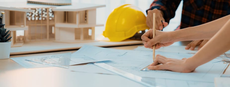 Cropped image of cooperative architect team decide and work together on meeting table with house model, safety helmet and architectural plan scatter around. Closeup. Focus on hand. Burgeoning.