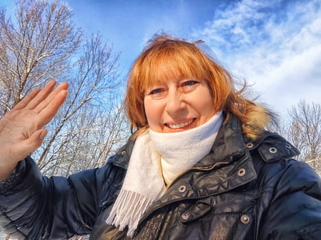 A cheerful middle aged woman in a winter coat and scarf taking selfie on nature outdoors and cold, snowy background with blue sky and white clouds