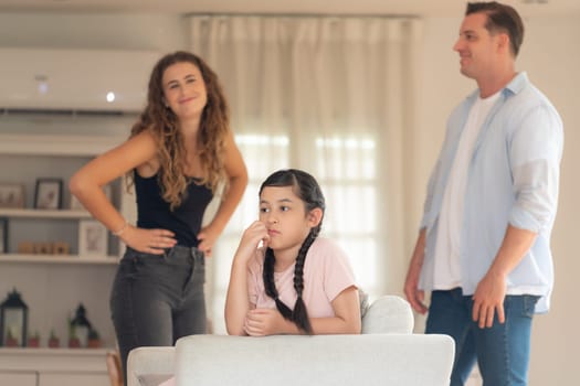 Annoyed and unhappy young girl sitting on sofa trapped in middle of tension by her parent argument in living room. Unhealthy domestic lifestyle and traumatic childhood develop to depression.Synchronos