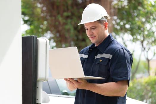 Qualified technician working on home EV charging station installation, making troubleshooting and configuration setup on charging system with laptop for EV at home. Synchronos