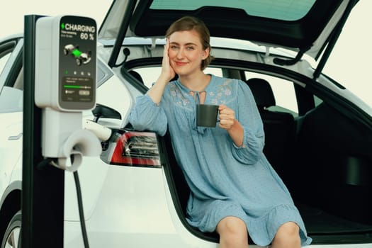Holiday road trip vacation with young woman sitting in the car trunk with coffee while recharging electric vehicle with alternative energy. Environmental friendly travel wit EV car. Perpetual