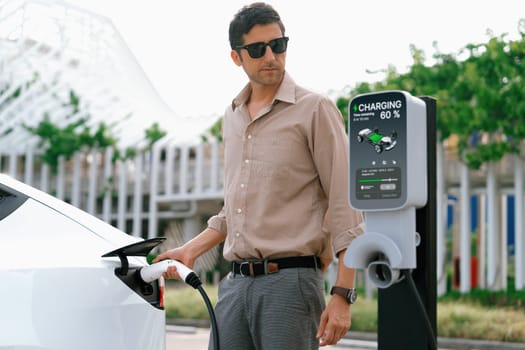 Young man put EV charger to recharge electric car's battery from charging station in city commercial parking lot. Rechargeable EV car for sustainable environmental friendly urban travel. Expedient