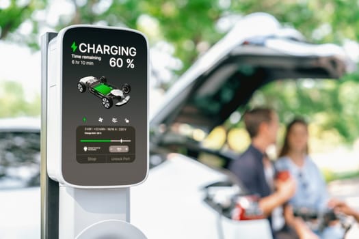 Focused EV charging station monitor display battery status for electric car on blurred background of couple sitting on car's trunk while recharging battery during natural travel and holiday. Exalt