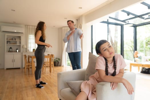 Annoyed and unhappy young girl sitting on sofa trapped in middle of tension by her parent argument in living room. Unhealthy domestic lifestyle and traumatic childhood develop to depression.Synchronos