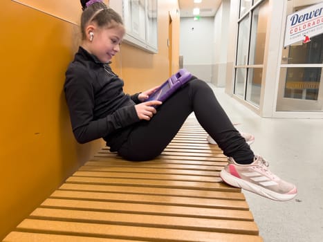 Denver, Colorado, USA-February 23, 2024- Nestled in a quiet hallway, this dedicated young figure skater finds a moment of tranquility, engrossed in her tablet as she mentally prepares for her upcoming performance.