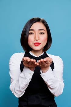 Smiling young woman receptionist blowing kiss with hands and looking at camera with positive facial expression. Cafe cheerful asian waitress showing love portrait on blue background
