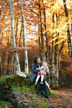 Mom with a little girl in her arms sits on a stump in the autumn forest. High quality photo